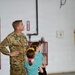 A U.S. military service member in OCP (Operational Camouflage Pattern) uniform stands in what appears to be an aircraft hangar alongside two young girls, one of whom is wearing a 'Happy Birthday' tiara. All three are looking upward, likely at an aircraft or display above them. The setting suggests a family day or open house event at a military installation.