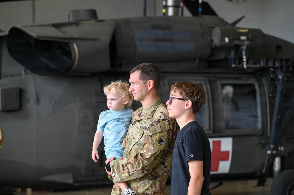 A U.S. Army soldier in OCP (Operational Camouflage Pattern) uniform holds a young toddler while standing next to an older child, approximately 10-12 years old. They are positioned in front of a military helicopter inside a hangar. The helicopter displays a red cross marking, indicating it is a medical/medevac aircraft.