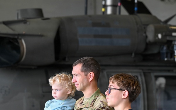 A U.S. Army soldier in OCP (Operational Camouflage Pattern) uniform holds a young toddler while standing next to an older child, approximately 10-12 years old. They are positioned in front of a military helicopter inside a hangar. The helicopter displays a red cross marking, indicating it is a medical/medevac aircraft.