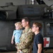 A U.S. Army soldier in OCP (Operational Camouflage Pattern) uniform holds a young toddler while standing next to an older child, approximately 10-12 years old. They are positioned in front of a military helicopter inside a hangar. The helicopter displays a red cross marking, indicating it is a medical/medevac aircraft.