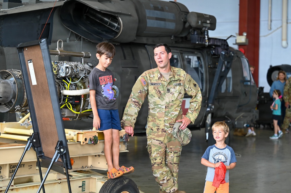 A U.S. Army soldier in OCP (Operational Camouflage Pattern) uniform is interacting with two young children inside a military helicopter hangar. The soldier, whose name tape reads 'HANCOCK,' appears to be explaining or demonstrating equipment near a disassembled helicopter engine. The setting appears to be a family day or open house event at a military installation.
