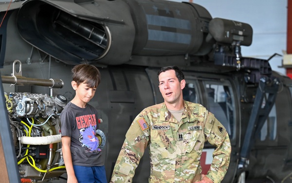 A U.S. Army soldier in OCP (Operational Camouflage Pattern) uniform is interacting with two young children inside a military helicopter hangar. The soldier, whose name tape reads 'HANCOCK,' appears to be explaining or demonstrating equipment near a disassembled helicopter engine. The setting appears to be a family day or open house event at a military installation.
