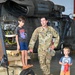 A U.S. Army soldier in OCP (Operational Camouflage Pattern) uniform is interacting with two young children inside a military helicopter hangar. The soldier, whose name tape reads 'HANCOCK,' appears to be explaining or demonstrating equipment near a disassembled helicopter engine. The setting appears to be a family day or open house event at a military installation.