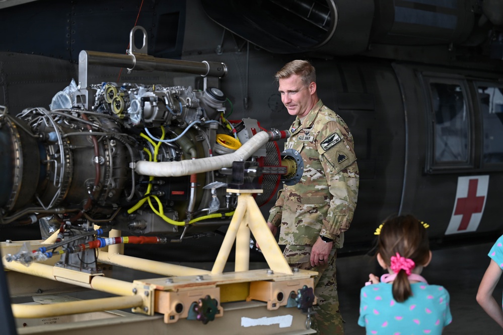 A U.S. Army soldier in OCP (Operational Camouflage Pattern) uniform is presenting or explaining a helicopter turbine engine on a maintenance stand to civilian visitors, including a young girl. The setting appears to be a military open house or public outreach event, indicated by the informal civilian presence. In the background, a military helicopter with a red cross marking (medevac/medical) is visible.