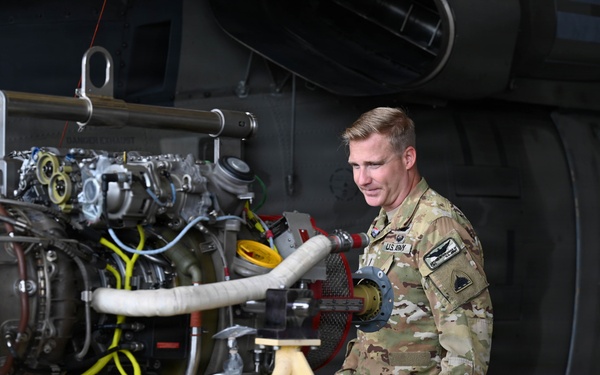 A U.S. Army soldier in OCP (Operational Camouflage Pattern) uniform is presenting or explaining a helicopter turbine engine on a maintenance stand to civilian visitors, including a young girl. The setting appears to be a military open house or public outreach event, indicated by the informal civilian presence. In the background, a military helicopter with a red cross marking (medevac/medical) is visible.