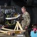 A U.S. Army soldier in OCP (Operational Camouflage Pattern) uniform is presenting or explaining a helicopter turbine engine on a maintenance stand to civilian visitors, including a young girl. The setting appears to be a military open house or public outreach event, indicated by the informal civilian presence. In the background, a military helicopter with a red cross marking (medevac/medical) is visible.