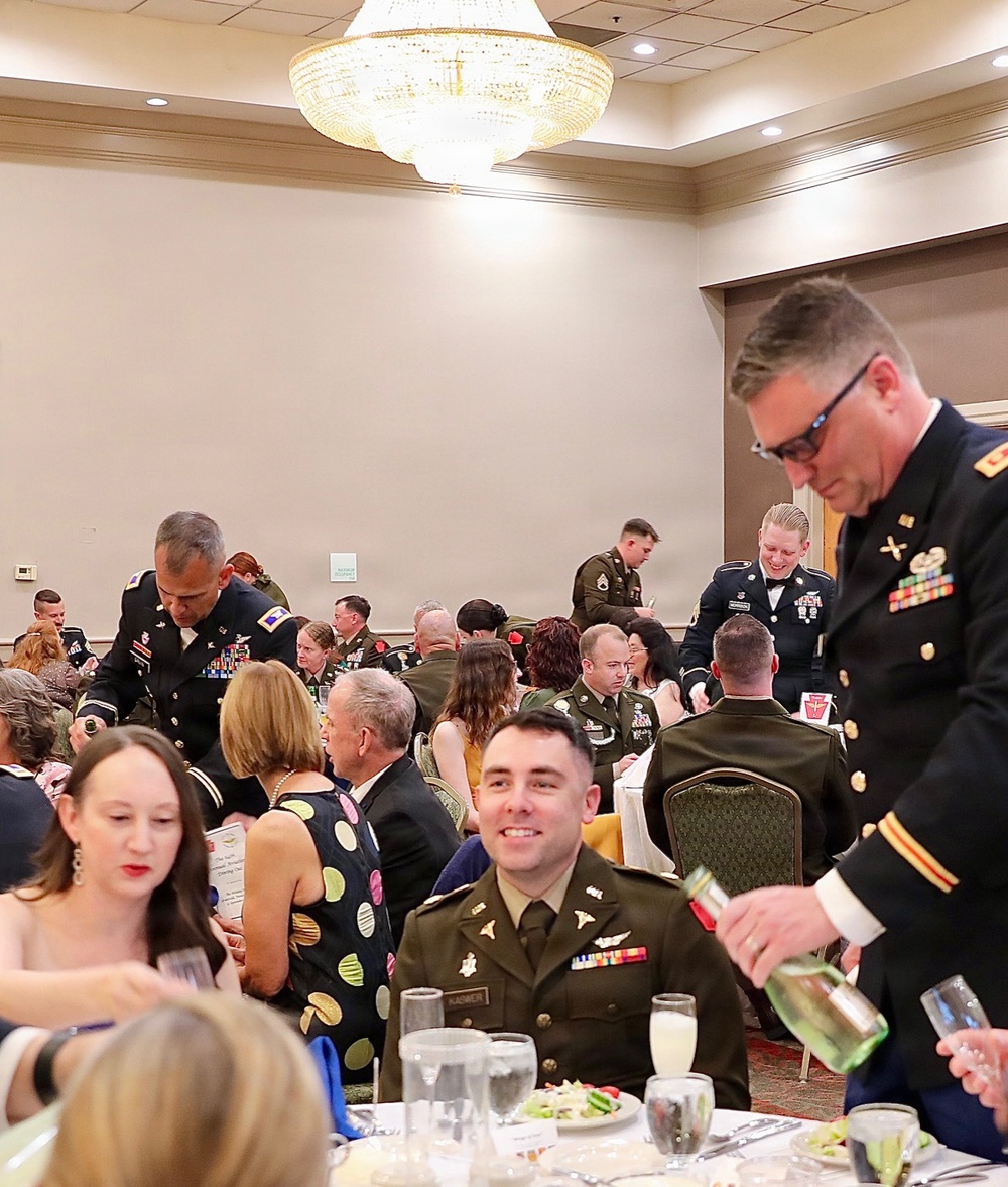 A formal military dining event, likely a military ball or formal dinner, taking place in a hotel ballroom. Multiple U.S. Army personnel in dress uniforms (Army Service Uniform/Class A) are seated and mingling at round tables alongside civilian guests in formal attire. The setting features a large crystal chandelier, white tablecloths, and formal place settings.