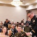 A formal military dining event, likely a military ball or formal dinner, taking place in a hotel ballroom. Multiple U.S. Army personnel in dress uniforms (Army Service Uniform/Class A) are seated and mingling at round tables alongside civilian guests in formal attire. The setting features a large crystal chandelier, white tablecloths, and formal place settings.
