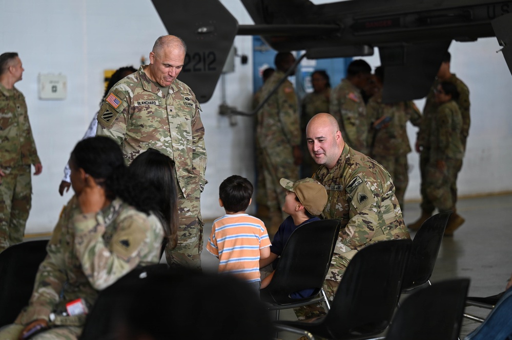 The image shows a military gathering inside what appears to be an aircraft hangar, with U.S. Army soldiers in OCP (Operational Camouflage Pattern) uniforms interacting with civilians including children. A senior officer identified by the name tape 'BLANCHARD' and a general officer star is visible speaking with a soldier who is engaging with two young boys. The setting suggests a family day, unit ceremony, or pre/post-deployment event.