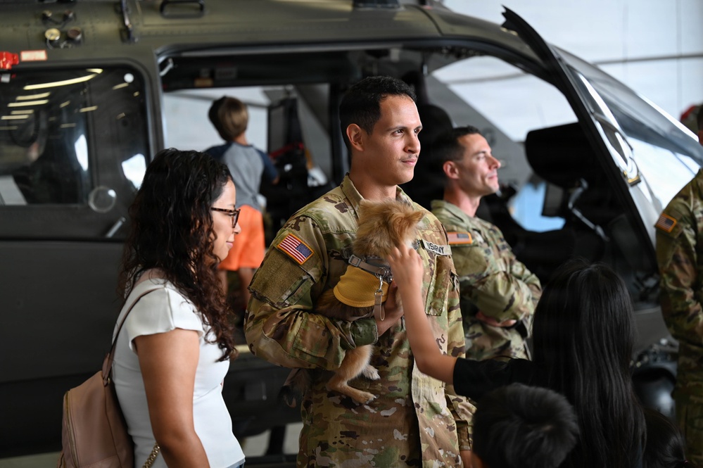 A U.S. Army soldier in OCP (Operational Camouflage Pattern) uniform holds a small dog while standing next to a civilian woman in what appears to be a military hangar. A military helicopter is visible in the background along with other soldiers and civilians. The scene appears to be a family day event or homecoming ceremony at a military aviation unit.