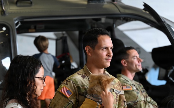 A U.S. Army soldier in OCP (Operational Camouflage Pattern) uniform holds a small dog while standing next to a civilian woman in what appears to be a military hangar. A military helicopter is visible in the background along with other soldiers and civilians. The scene appears to be a family day event or homecoming ceremony at a military aviation unit.