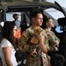 A U.S. Army soldier in OCP (Operational Camouflage Pattern) uniform holds a small dog while standing next to a civilian woman in what appears to be a military hangar. A military helicopter is visible in the background along with other soldiers and civilians. The scene appears to be a family day event or homecoming ceremony at a military aviation unit.