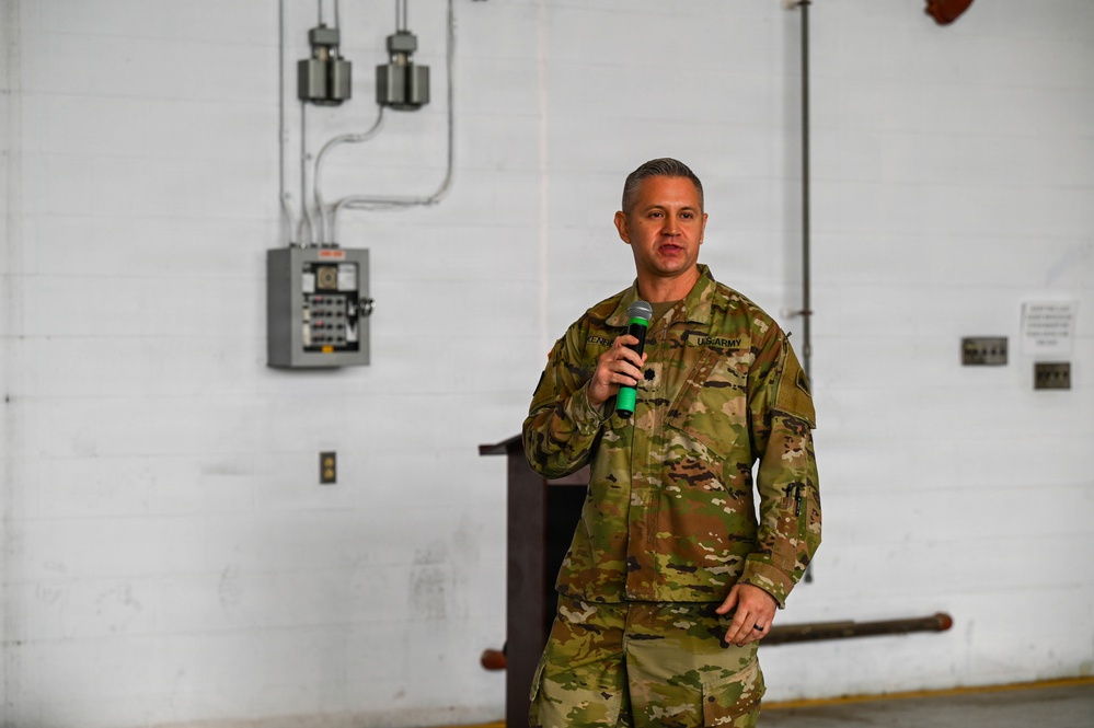 A U.S. Army soldier in Operational Camouflage Pattern (OCP) uniform is speaking into a green-handled microphone, apparently addressing an audience. The setting appears to be an industrial or warehouse-type facility with exposed electrical conduits and a circuit breaker panel visible on the white brick wall behind him. A wooden podium is partially visible in the background.