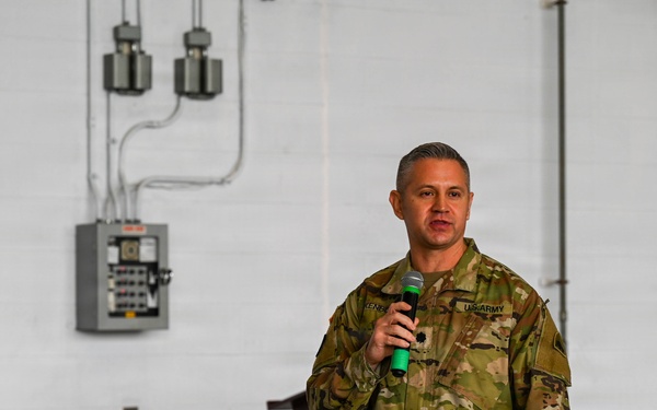 A U.S. Army soldier in Operational Camouflage Pattern (OCP) uniform is speaking into a green-handled microphone, apparently addressing an audience. The setting appears to be an industrial or warehouse-type facility with exposed electrical conduits and a circuit breaker panel visible on the white brick wall behind him. A wooden podium is partially visible in the background.