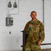 A U.S. Army soldier in Operational Camouflage Pattern (OCP) uniform is speaking into a green-handled microphone, apparently addressing an audience. The setting appears to be an industrial or warehouse-type facility with exposed electrical conduits and a circuit breaker panel visible on the white brick wall behind him. A wooden podium is partially visible in the background.