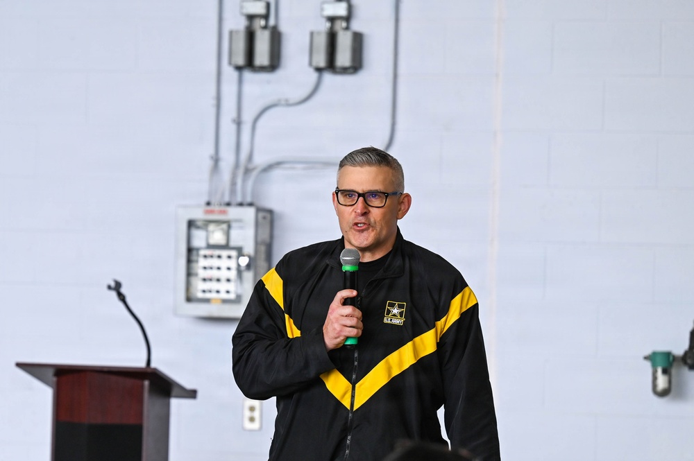 A middle-aged man wearing a U.S. Army branded black and yellow athletic jacket is speaking into a handheld microphone. He appears to be addressing an audience in what looks like a gymnasium or utility room setting. A wooden podium is visible to his left, suggesting a formal or semi-formal presentation.