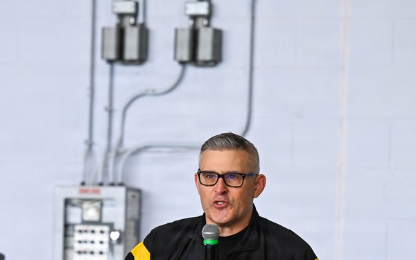A middle-aged man wearing a U.S. Army branded black and yellow athletic jacket is speaking into a handheld microphone. He appears to be addressing an audience in what looks like a gymnasium or utility room setting. A wooden podium is visible to his left, suggesting a formal or semi-formal presentation.