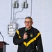 A middle-aged man wearing a U.S. Army branded black and yellow athletic jacket is speaking into a handheld microphone. He appears to be addressing an audience in what looks like a gymnasium or utility room setting. A wooden podium is visible to his left, suggesting a formal or semi-formal presentation.