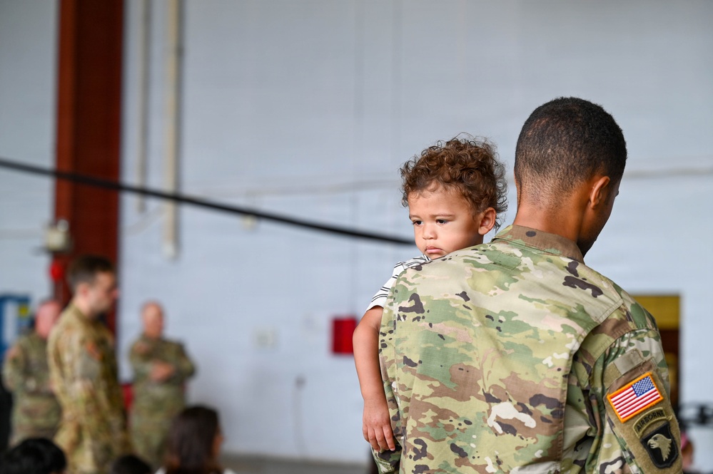 A U.S. Army soldier in OCP (Operational Camouflage Pattern) uniform is holding a young toddler with curly hair during what appears to be a military ceremony or formation inside a hangar. The soldier's right shoulder displays a U.S. flag patch and a 101st Airborne Division patch. Several other soldiers in uniform are visible in the blurred background.