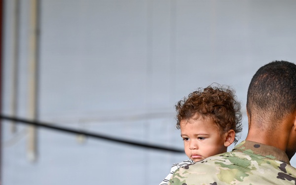 A U.S. Army soldier in OCP (Operational Camouflage Pattern) uniform is holding a young toddler with curly hair during what appears to be a military ceremony or formation inside a hangar. The soldier's right shoulder displays a U.S. flag patch and a 101st Airborne Division patch. Several other soldiers in uniform are visible in the blurred background.