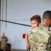 A U.S. Army soldier in OCP (Operational Camouflage Pattern) uniform is holding a young toddler with curly hair during what appears to be a military ceremony or formation inside a hangar. The soldier's right shoulder displays a U.S. flag patch and a 101st Airborne Division patch. Several other soldiers in uniform are visible in the blurred background.