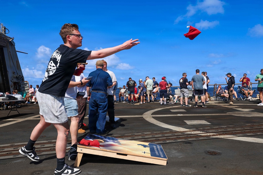 A group of people are playing cornhole on what appears to be the deck of a military naval vessel, identifiable by the helicopter landing pad markings on the deck. A young man in a black graphic t-shirt is mid-throw, releasing a red bean bag toward the cornhole board decorated with an American flag design. Multiple spectators and other players are gathered in the background against a bright blue sky over open ocean.