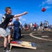 A group of people are playing cornhole on what appears to be the deck of a military naval vessel, identifiable by the helicopter landing pad markings on the deck. A young man in a black graphic t-shirt is mid-throw, releasing a red bean bag toward the cornhole board decorated with an American flag design. Multiple spectators and other players are gathered in the background against a bright blue sky over open ocean.