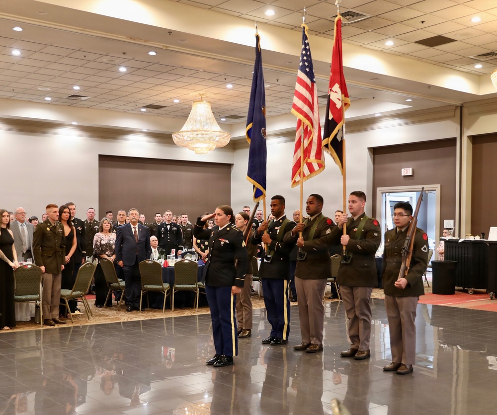 A military color guard ceremony is taking place in a formal banquet hall, with uniformed soldiers presenting flags including the American flag and what appears to be a unit or organizational flag. A female soldier in the foreground is rendering a salute while other soldiers hold the colors and rifles. Attendees in both military dress uniforms and civilian attire stand at attention in the background.