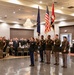 A military color guard ceremony is taking place in a formal banquet hall, with uniformed soldiers presenting flags including the American flag and what appears to be a unit or organizational flag. A female soldier in the foreground is rendering a salute while other soldiers hold the colors and rifles. Attendees in both military dress uniforms and civilian attire stand at attention in the background.