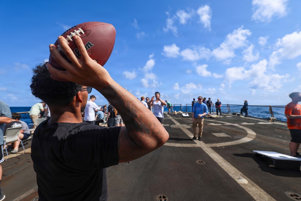A man with tattoos on his arm is preparing to throw a Wilson football on what appears to be the deck of a naval vessel at sea. Multiple people are gathered on the deck engaging in recreational activities, with the ocean and partly cloudy sky visible in the background. The circular marking on the deck suggests this may be a helicopter landing pad being used as a recreational space.