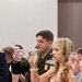 A formal military banquet or ball event where attendees are raising glasses in a toast. A male officer in U.S. Army dress green uniform with multiple service ribbons, badges, and rank insignia stands prominently alongside a woman in a floral evening dress. The setting appears to be a hotel ballroom with dining tables visible in the foreground.