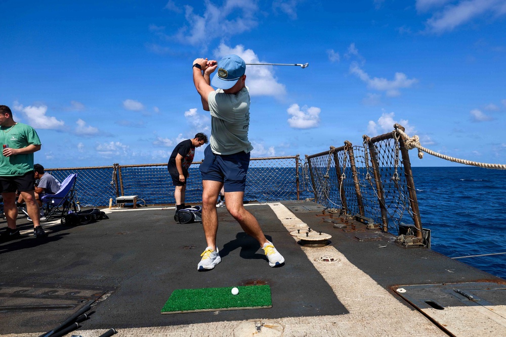 A person is playing golf on the deck of a naval vessel at sea, having just completed a swing from a small artificial turf mat. Several other individuals are visible in the background on the ship's deck, which is surrounded by safety netting and railings with open ocean visible beyond. The scene depicts an informal recreational activity taking place aboard a military ship.