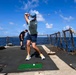 A person is playing golf on the deck of a naval vessel at sea, having just completed a swing from a small artificial turf mat. Several other individuals are visible in the background on the ship's deck, which is surrounded by safety netting and railings with open ocean visible beyond. The scene depicts an informal recreational activity taking place aboard a military ship.