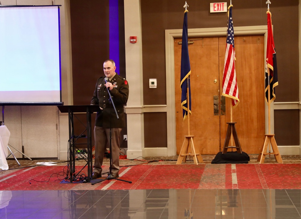 A military officer in dress uniform is speaking at a podium with a microphone during what appears to be a formal ceremony or event held in a banquet or conference hall. Three flags are displayed on wooden stands in the background, including the United States flag and what appears to be a state flag. A projection screen is visible to the left, suggesting a presentation is part of the event.