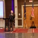 A military officer in dress uniform is speaking at a podium with a microphone during what appears to be a formal ceremony or event held in a banquet or conference hall. Three flags are displayed on wooden stands in the background, including the United States flag and what appears to be a state flag. A projection screen is visible to the left, suggesting a presentation is part of the event.