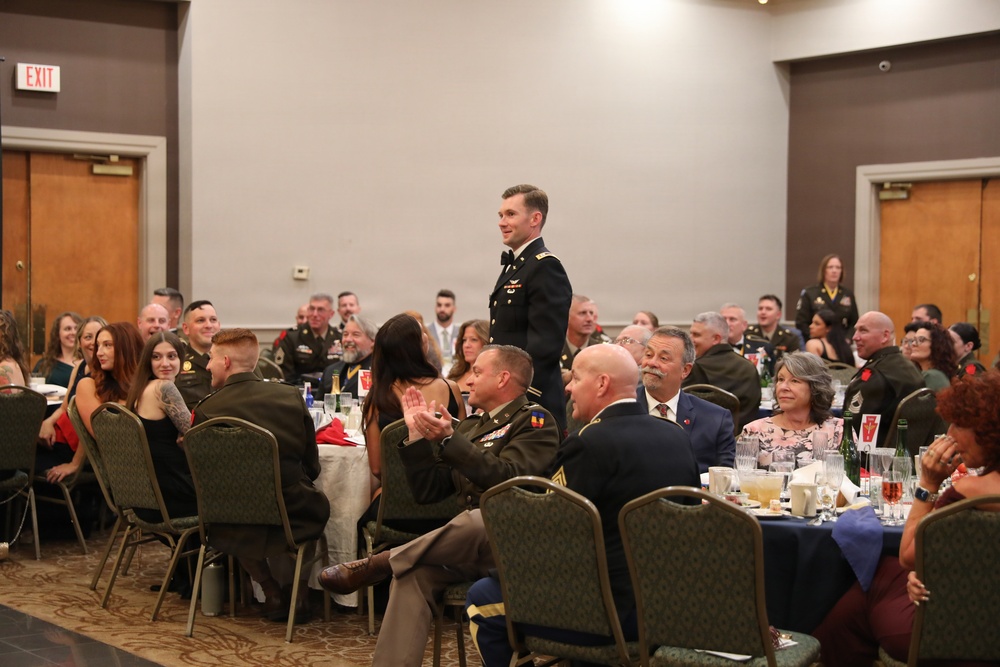 A formal military banquet or dining-in event is taking place in a banquet hall, with numerous attendees in U.S. Army dress uniforms seated at round tables. One uniformed soldier is standing, appearing to address or acknowledge the assembled group while others look on and applaud. The event appears to be a formal recognition or awards ceremony based on the attire and setting.