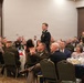 A formal military banquet or dining-in event is taking place in a banquet hall, with numerous attendees in U.S. Army dress uniforms seated at round tables. One uniformed soldier is standing, appearing to address or acknowledge the assembled group while others look on and applaud. The event appears to be a formal recognition or awards ceremony based on the attire and setting.