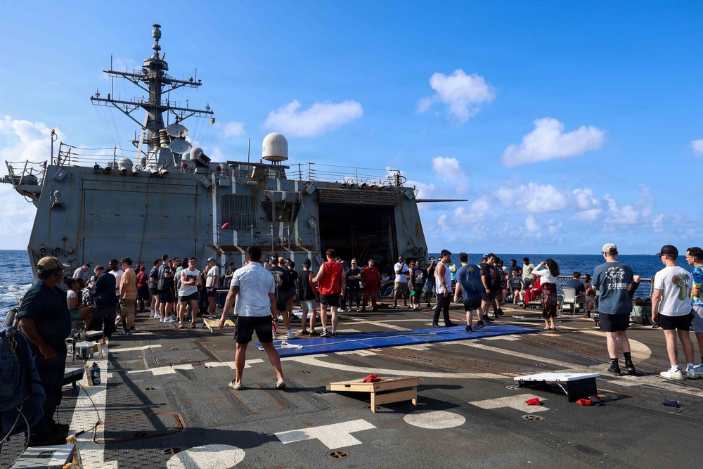 A large group of military personnel are gathered on the flight deck or well deck of a U.S. Navy warship, appearing to be participating in a morale or recreation day at sea. The ship's superstructure, radar domes, and communications equipment are clearly visible in the background. Personnel are dressed in casual civilian clothing, suggesting an off-duty or liberty event.