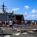 A large group of military personnel are gathered on the flight deck or well deck of a U.S. Navy warship, appearing to be participating in a morale or recreation day at sea. The ship's superstructure, radar domes, and communications equipment are clearly visible in the background. Personnel are dressed in casual civilian clothing, suggesting an off-duty or liberty event.