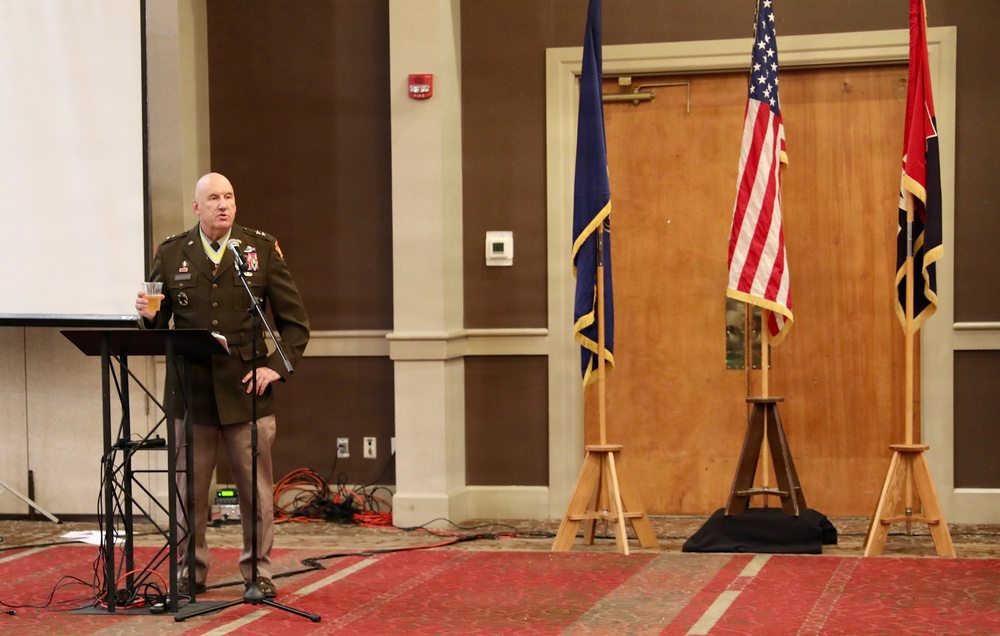 A military officer in dress uniform stands at a podium delivering a speech at what appears to be a formal military ceremony or banquet. He is holding a glass, suggesting a toast may be in progress. Three flags are displayed on wooden stands behind him, including the United States flag and what appears to be a state flag.