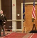 A military officer in dress uniform stands at a podium delivering a speech at what appears to be a formal military ceremony or banquet. He is holding a glass, suggesting a toast may be in progress. Three flags are displayed on wooden stands behind him, including the United States flag and what appears to be a state flag.