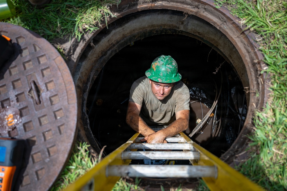A male worker wearing a green hard hat is climbing up a ladder out of an open manhole or underground utility access point. The image is taken from above, looking down at the worker emerging from the confined space. Safety and monitoring equipment is visible at ground level beside the opening.