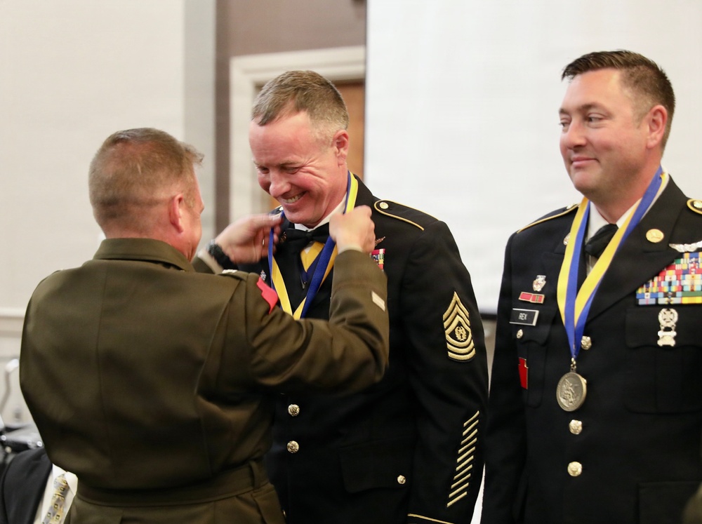 A military medal presentation ceremony is taking place indoors. A Marine Corps officer (facing away) is placing a medal on a ribbon around the neck of a smiling Army Sergeant Major, while a second Army soldier already wearing an identical medal looks on. The atmosphere appears celebratory and informal in tone despite the formal military setting.