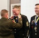 A military medal presentation ceremony is taking place indoors. A Marine Corps officer (facing away) is placing a medal on a ribbon around the neck of a smiling Army Sergeant Major, while a second Army soldier already wearing an identical medal looks on. The atmosphere appears celebratory and informal in tone despite the formal military setting.
