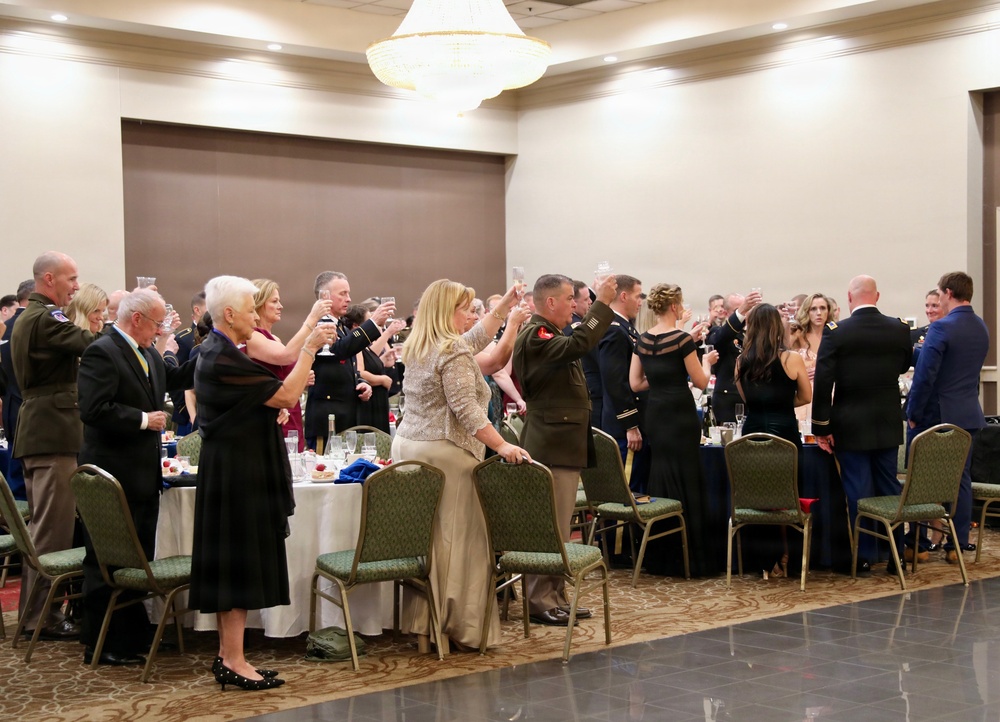 A formal military banquet or ball is taking place in a hotel ballroom, with attendees in military dress uniforms and formal civilian attire raising glasses in a toast. The event appears to be a structured ceremonial dinner, likely a military ball or gala, with round banquet tables set with white linens and glassware. Multiple military personnel from what appears to be the U.S. Army and/or Marine Corps are present alongside civilian guests.