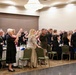 A formal military banquet or ball is taking place in a hotel ballroom, with attendees in military dress uniforms and formal civilian attire raising glasses in a toast. The event appears to be a structured ceremonial dinner, likely a military ball or gala, with round banquet tables set with white linens and glassware. Multiple military personnel from what appears to be the U.S. Army and/or Marine Corps are present alongside civilian guests.