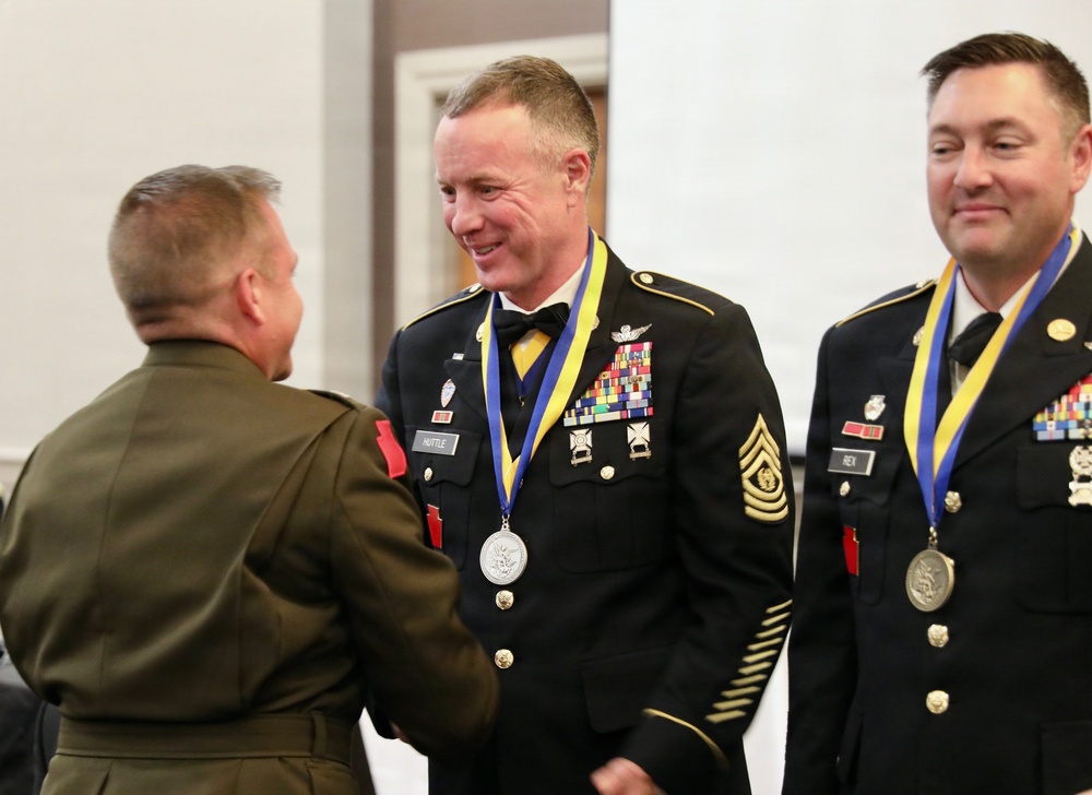 A military awards ceremony is taking place where two U.S. Army soldiers in dress uniforms are receiving medallions on blue and yellow ribbons. A Marine officer in service uniform is shaking hands with one of the soldiers, identified by a name tag as 'HUTTLE', who holds a senior NCO rank (Sergeant Major or higher based on chevrons). Both Army recipients wear extensive medal racks and formal dress uniforms with bow ties.