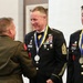 A military awards ceremony is taking place where two U.S. Army soldiers in dress uniforms are receiving medallions on blue and yellow ribbons. A Marine officer in service uniform is shaking hands with one of the soldiers, identified by a name tag as 'HUTTLE', who holds a senior NCO rank (Sergeant Major or higher based on chevrons). Both Army recipients wear extensive medal racks and formal dress uniforms with bow ties.