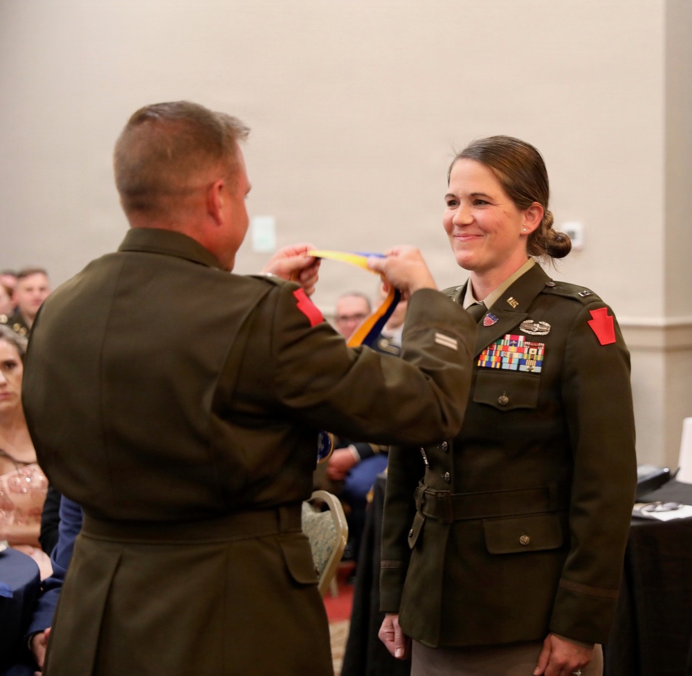 A military award ceremony is taking place in what appears to be a banquet or conference hall. A male officer is placing a medal or decoration on a ribbon around the neck of a female officer, who is smiling. Both individuals are wearing U.S. Army dress green uniforms with multiple decorations and insignia.