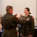 A military award ceremony is taking place in what appears to be a banquet or conference hall. A male officer is placing a medal or decoration on a ribbon around the neck of a female officer, who is smiling. Both individuals are wearing U.S. Army dress green uniforms with multiple decorations and insignia.